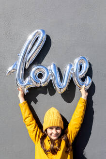 Ein glückliches junges Mädchen mit gelbem Hut und Mantel hebt einen silbern glänzenden Liebesballon gegen eine graue Betonmauer und teilt einen freudigen Moment - ADSF55247
