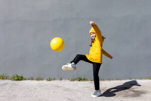 Lebhafte junge Frau in Gelb spielt mit einem Ball vor einer grauen Wand - ADSF55249