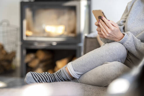 Young Woman sitting on the sofa in a clean modern home with his phone browsing with a warm fire in the background - ISF26938