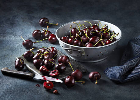 A bowl of fresh cherries on a textured dark surface with a few cherries scattered around and a cherry with a bite taken out of it - ISF26993