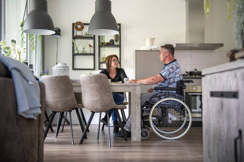 Eine Frau bereitet das Mittagessen für ihren Mann vor, der in seinem Rollstuhl am Tisch sitzt - ISF27119