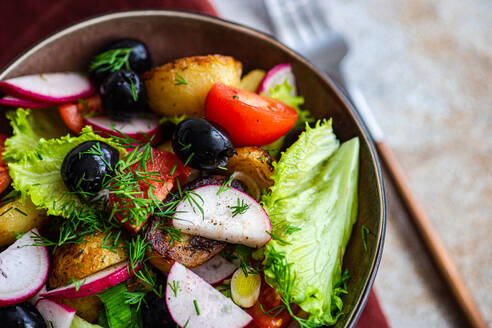 A close-up of a delicious Mediterranean salad bowl, with golden potatoes, ripe cherry tomatoes, black olives, spring onions, radishes, and lettuce, tastefully garnished with dill - ADSF55296