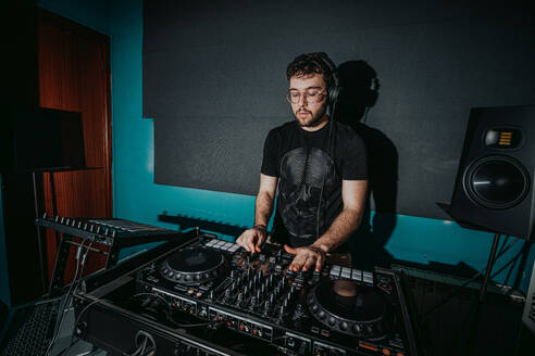 A focused male DJ mixes tracks on a professional sound mixer, with headphones and studio monitors in a dark setting - ADSF55327