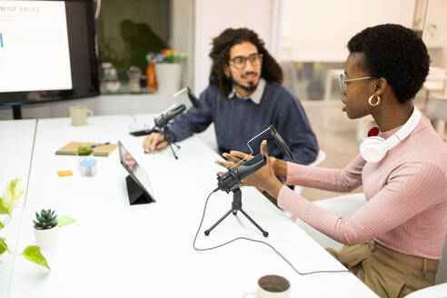 A Black woman adjusts equipment while a man with curly hair, likely of Middle Eastern or Hispanic ethnicity, looks at her in a podcasting setup. - ADSF55443