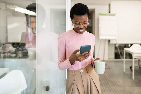 Cheerful African American woman looking at her phone and holding a coffee mug, standing in an office environment. - ADSF55450