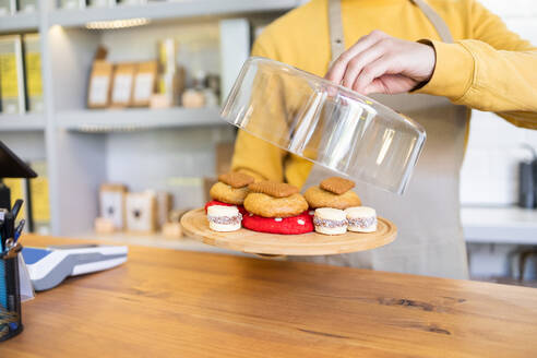 A person in a yellow apron reveals a variety of freshly baked cookies on a wooden plate in a contemporary bakery setting. - ADSF55463