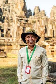 A cheerful local male guide wearing a safari hat and a lanyard smiles in front of the ancient Angkor Wat Hindu temple ruins in Siem Reap, Cambodia. - ADSF55534