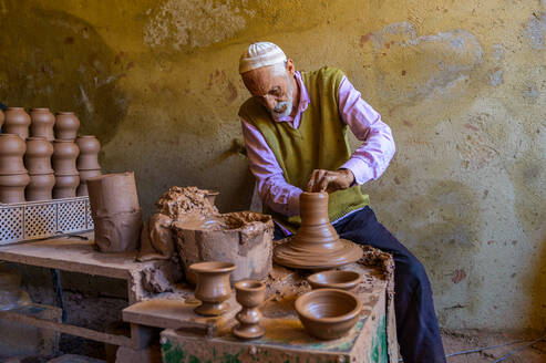 An experienced artisan shapes clay on a pottery wheel, surrounded by handmade pots in a rustic workshop. - ADSF55560