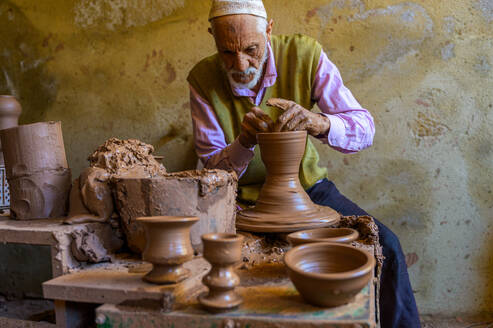 An elderly potter shapes clay on a wheel, creating traditional ceramics in a rustic workshop. - ADSF55561