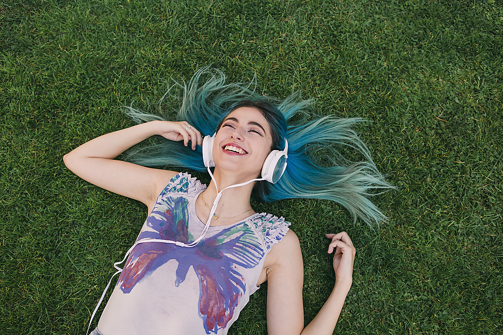 Glückliche, unbekümmerte junge Frau mit blauen Haaren, die mit Kopfhörern Musik hört und im Gras liegt