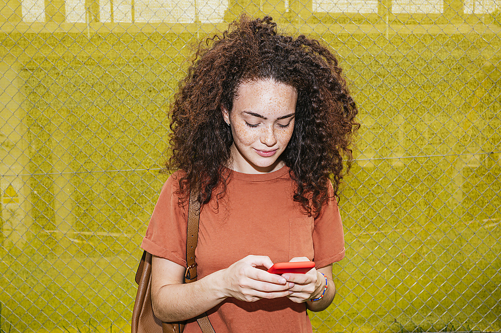 Junge Frau mit lockigem Haar, die mit ihrem Smartphone vor einem gelben Zaun Textnachrichten verschickt