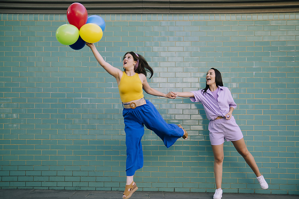 Woman catching friend holding multi colored balloons in front of turquoise wall